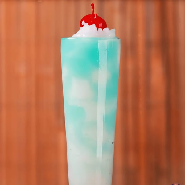 Tall glass of Caribbean blue drink with white foam and a red cherry on top.