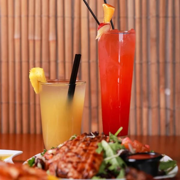 Two vibrant Caribbean cocktails and a plate of grilled meat with salad, against a bamboo background.