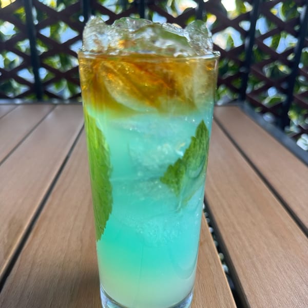 A vibrant blue and yellow layered cocktail with mint leaves and ice on a wooden table.